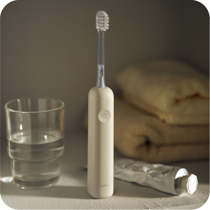 Beige electric toothbrush with a glass of water and tube of toothpaste on a towel