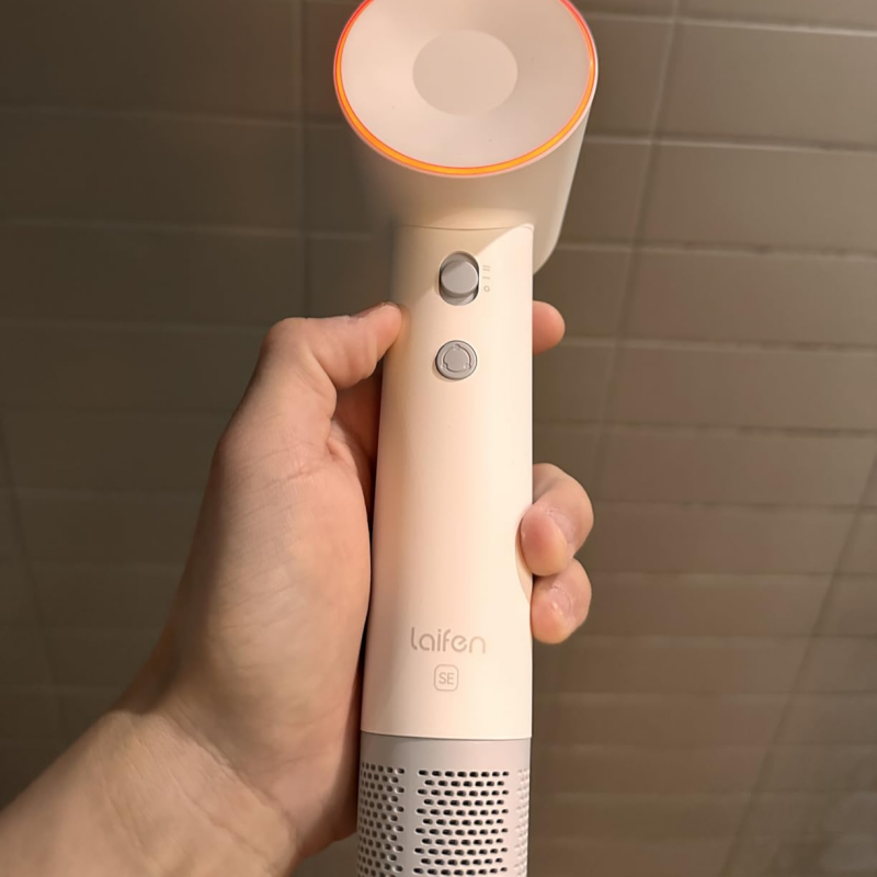 Hand holding a white handheld device with an orange accent, brand 'laifen' visible, against a tiled wall background.